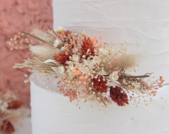 May include: A white tiered cake decorated with a dried floral arrangement. The arrangement features a mix of beige, orange, and white flowers, including small blooms and feathery accents. The cake has a textured, buttercream frosting.