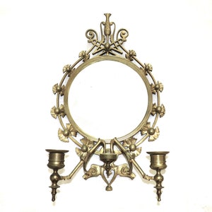 May include: A gold-toned wall mirror with ornate floral details and two candle holders on either side. The mirror is round and has a simple, plain glass surface.