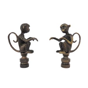 May include: Two bronze-colored monkey figurines. Each monkey is seated with arms outstretched, atop a decorative base. The monkeys have a tail that curves into a loop. The background is white.