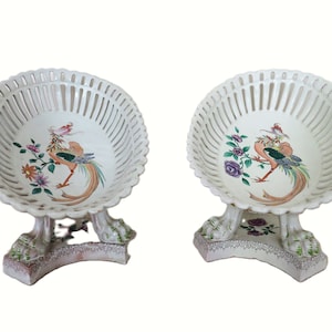 May include: Two white ceramic serving bowls with openwork rims and pedestal bases. Each bowl features a colorful bird and floral design. The bases have a decorative floral pattern. A classic piece for serving fruit or displaying decorative items.