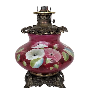 May include: A vintage oil lamp featuring a deep magenta glass base decorated with hand-painted white and pink morning glory flowers and green leaves. The lamp includes an ornate brass base and a decorative brass collar with a metal burner assembly.
