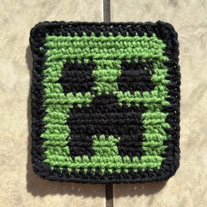 Pode incluir: Um quadrado de crochê verde e preto com um rosto de creeper preto no centro.