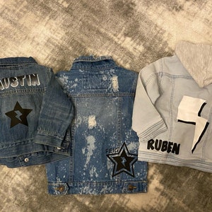 May include: Three denim jackets with personalised designs. The first jacket is dark blue with the name "Austin" and a black star with a lightning bolt. The second jacket is light blue with white paint splatters and the name "Feig" and a black star with a lightning bolt. The third jacket is light blue with a white lightning bolt and the name "Ruben".