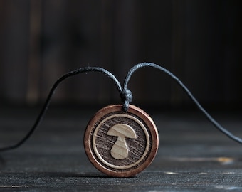 May include: A wooden pendant necklace featuring a mushroom design. The pendant is brown with a light brown mushroom carved into it. The necklace is made of black cord.