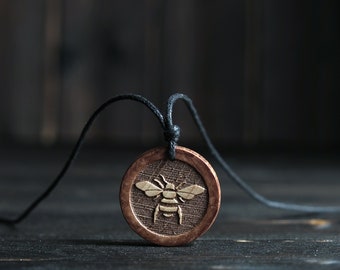 Puede incluir: Un collar con un colgante de madera y un cordón negro. El colgante es redondo y presenta una imagen tallada de una abeja.