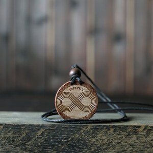 Op de afbeelding: Een houten ketting met een hanger in de vorm van een oneindigheidssymbool en het woord "INFINITY" erop gegraveerd. De hanger is bruin en het koord is zwart.