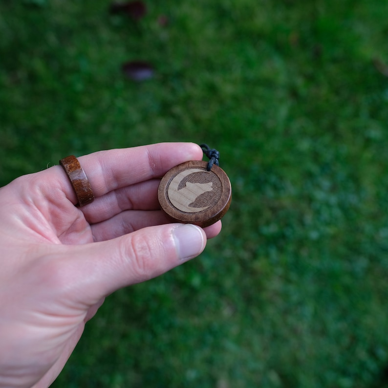 May include: A wooden pendant with a wolf silhouette carved into the wood. The pendant is on a black cord and is being held by a hand.
