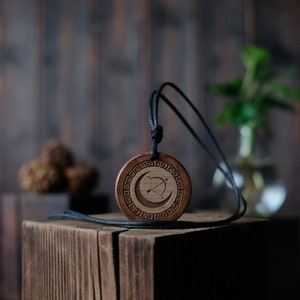 May include: A wooden pendant with a crescent moon and bow and arrow design, hanging from a black cord. The pendant is on a wooden surface.