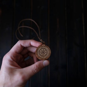 May include: A wooden pendant with a circular design featuring a Vegvisir symbol surrounded by runes. The pendant is on a brown leather cord.