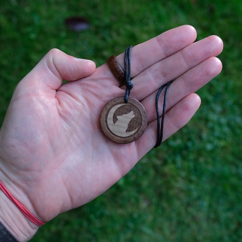 May include: A wooden pendant necklace with a black cord. The pendant features a carved wolf howling at the moon.