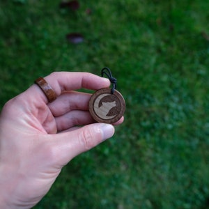 May include: A wooden pendant with a wolf howling at the moon design. The pendant is on a black cord and is being held by a hand.