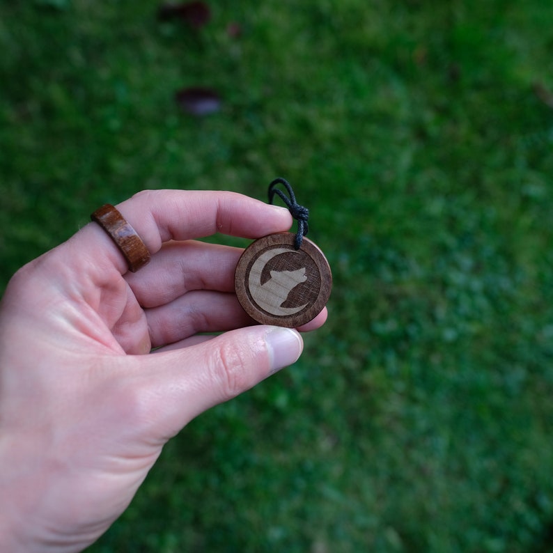May include: A wooden pendant with a wolf howling at the moon design. The pendant is on a black cord and is being held by a hand.
