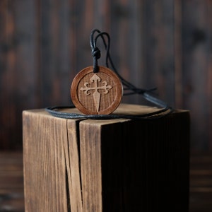 May include: A wooden pendant with a carved cross design hangs from a black cord. The pendant is round and has a light brown color. It is resting on a wooden block.