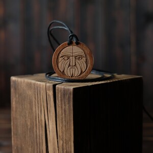 May include: A wooden pendant with a carved face design hangs on a black cord. The pendant is resting on a wooden block.