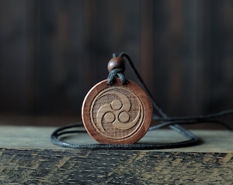 Puede incluir: Un colgante de madera con un cordón negro, que presenta un símbolo triquetra tallado. El colgante es marrón y tiene un acabado de madera natural.