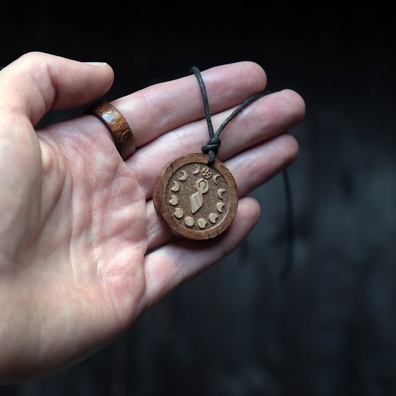 May include: A wooden pendant necklace with a brown cord. The pendant is round and features a carved design of a figure with a circle above its head, surrounded by a ring of crescent moons.