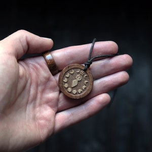 May include: A wooden pendant necklace with a brown cord. The pendant is round and features a carved design of a stylized figure with a circle above its head and eight crescent moons around the figure.