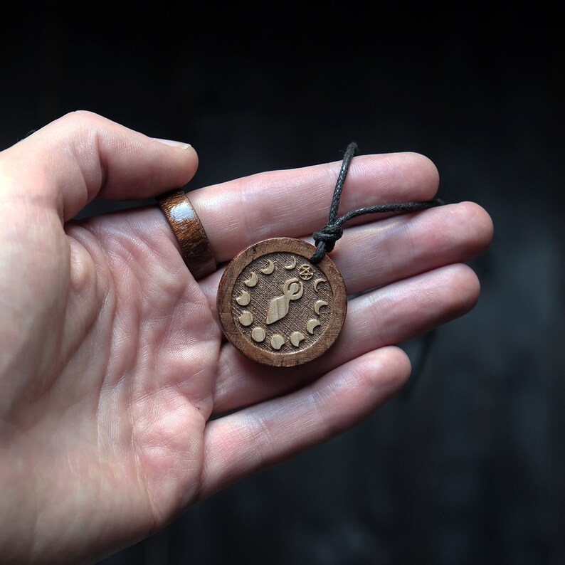 May include: A wooden pendant necklace with a brown cord. The pendant is round and features a carved design of a stylized figure with a circle above its head and eight crescent moons around the figure.