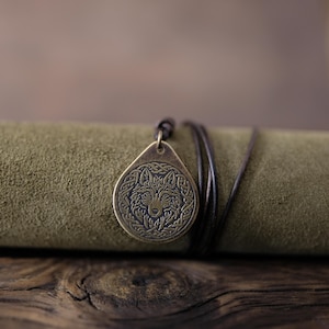 May include: A bronze pendant necklace with a wolf head design. The pendant is on a brown leather cord and is lying on a green suede surface.