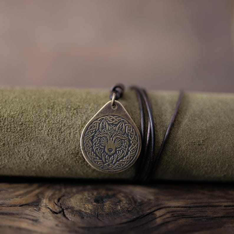 May include: A bronze pendant necklace with a wolf head design. The pendant is on a brown leather cord and is lying on a green suede surface.