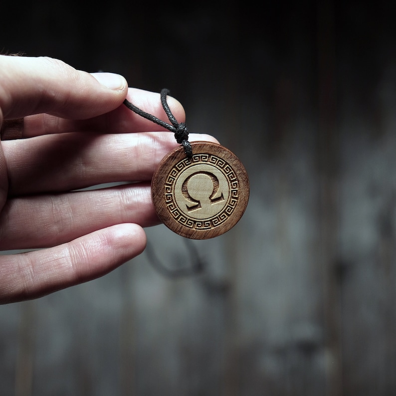 May include: A wooden pendant necklace with a black cord. The pendant is round and features a carved omega symbol surrounded by a Greek key pattern.