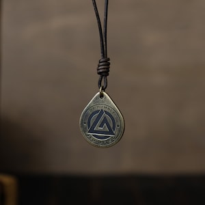 May include: A brown leather cord necklace with a brass pendant. The pendant is a teardrop shape with a black triangle and circle design. The text "BE HUMBLE JIU JITSU" is inscribed around the pendant.