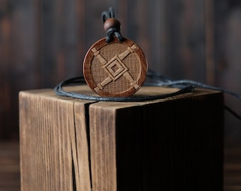 Puede incluir: Un collar con un colgante de madera con un diseño geométrico, que presenta un cuadrado dentro de un cuadrado más grande, en un cordón negro. El colgante está apoyado sobre una superficie de madera.