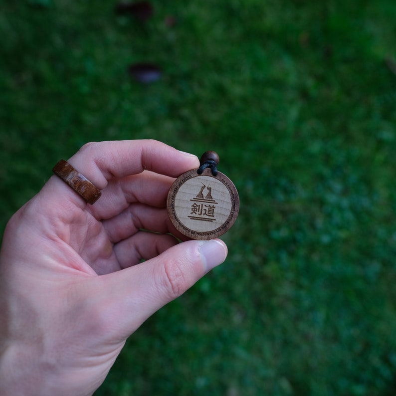 Puede incluir: Un colgante de madera con un cord&oacute;n negro, que presenta una imagen estilizada de dos figuras y el texto "剣道" en japon&eacute;s.