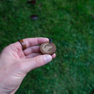 May include: A wooden pendant with a wolf silhouette design. The pendant is brown with a lighter wood inlay for the wolf. The pendant is hanging from a black cord.
