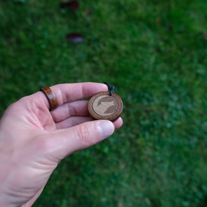 May include: A wooden pendant with a wolf silhouette design. The pendant is brown with a lighter wood inlay for the wolf. The pendant is hanging from a black cord.