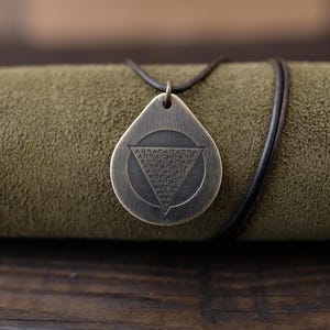 Op de afbeelding: Een bronzen hangerketting met een druppelvorm. De hanger heeft een cirkelvormig ontwerp met een driehoek erin, met het woord "ABRACADABRA" erop. De ketting heeft een donkerbruin leren koord.
