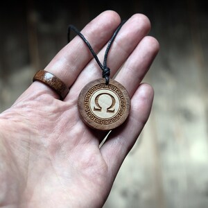 May include: A wooden pendant necklace with a black cord. The pendant is round and features a white omega symbol surrounded by a decorative border.