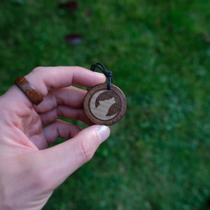 May include: A wooden pendant with a wolf howling at the moon design. The pendant is brown with a black cord.