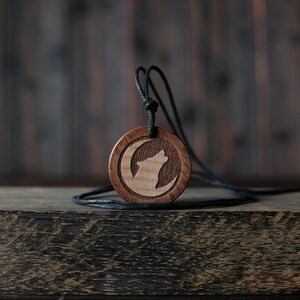May include: A wooden pendant necklace with a black cord. The pendant is round and features a carved wolf howling at the moon.