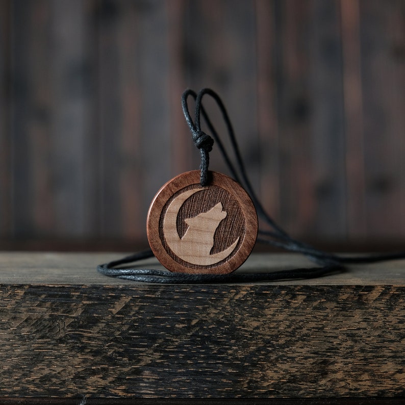 May include: A wooden pendant necklace with a black cord. The pendant is round and features a carved wolf howling at the moon.