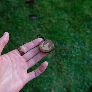 May include: A wooden pendant with a wolf howling at the moon design. The pendant is brown with a lighter wood inlay for the wolf and moon.