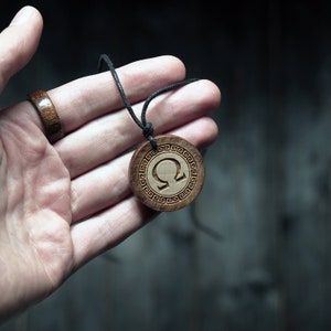 May include: A wooden pendant necklace with a black cord. The pendant is round and features a carved omega symbol surrounded by a Greek key pattern.