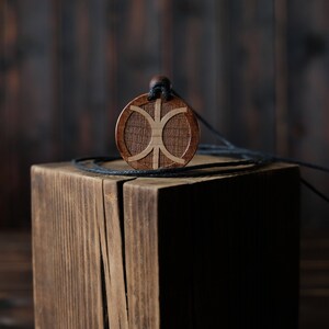 May include: A wooden pendant with a geometric design, hanging on a black cord, resting on a wooden block.