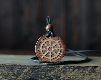 May include: A round, wooden pendant necklace featuring a carved Dharma wheel design. The pendant is brown with a lighter-colored wheel design. It hangs from a black cord with a wooden bead. The necklace rests on a wooden surface.