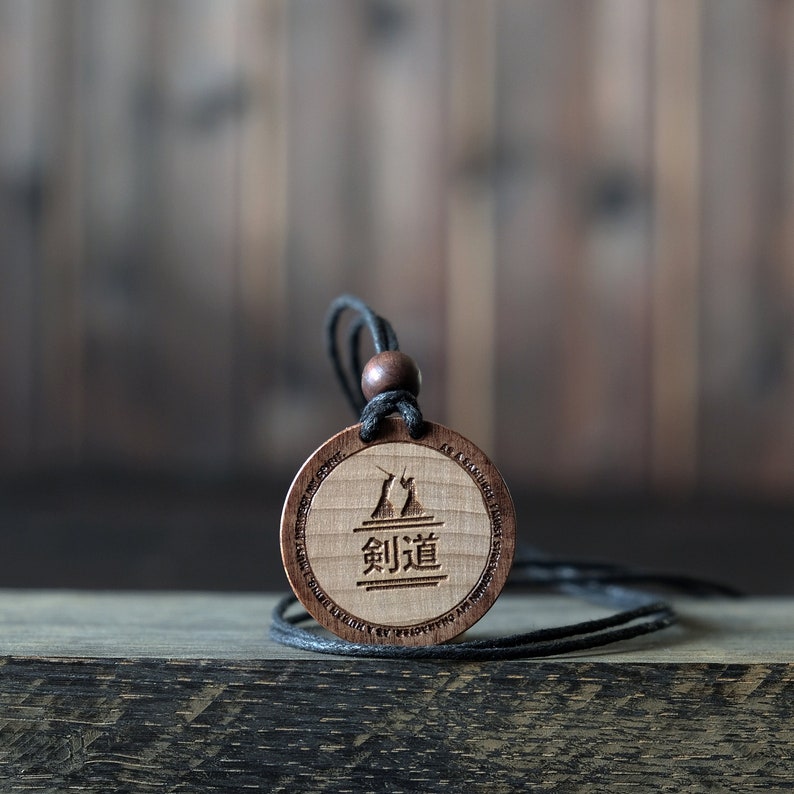 Puede incluir: Un collar con un colgante de madera y un cord&oacute;n negro. El colgante es redondo y presenta una imagen estilizada de dos figuras en una pose de esgrima con la palabra japonesa "剣道" (kendo) inscrita debajo.