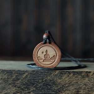 Collana/pendente Buddha. Realizzato in legno massello di sicomoro. Forma rotonda. Colore marrone. Cordoncino di cotone regolabile. Personalizzazione del testo.