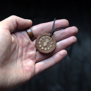 May include: A wooden pendant necklace with a brown cord. The pendant is round and features a carved design of a woman standing with a crescent moon above her head. The pendant is surrounded by a circle of smaller crescent moons.