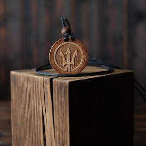May include: A round wooden pendant necklace with a carved trident design. The pendant is brown and hangs from a black cord with a wooden bead. The necklace is displayed on a wooden block against a dark background.