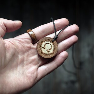May include: A wooden pendant necklace with a circular design featuring a Greek letter Q surrounded by a decorative border. The pendant is attached to a black cord.
