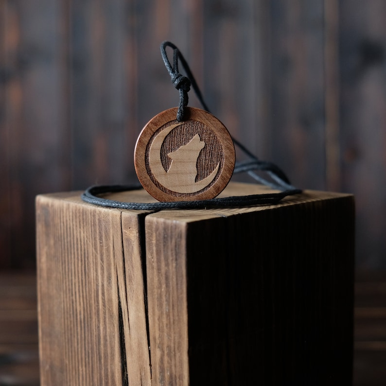 May include: A wooden pendant necklace with a wolf howling at the moon design. The pendant is brown and the cord is black.