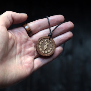 May include: A wooden pendant necklace with a brown cord. The pendant is round and features a carved design of a woman with a circle above her head and a crescent moon on each side. The pendant is surrounded by a circle of smaller crescent moons.