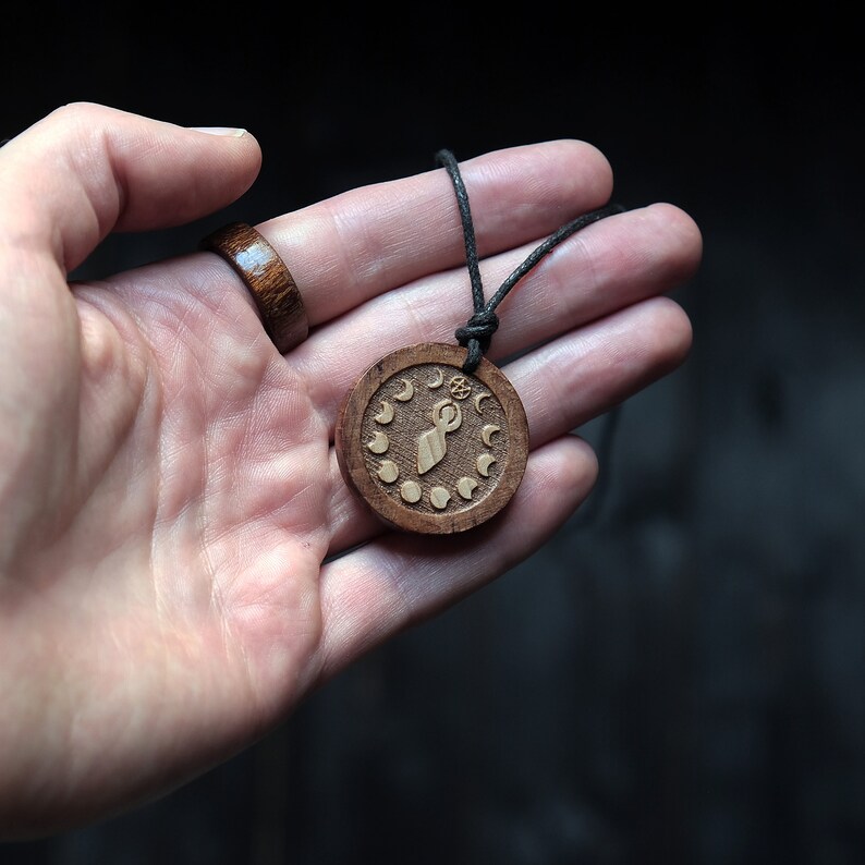 May include: A wooden pendant necklace with a brown cord. The pendant is round and features a carved design of a woman with a circle above her head and a crescent moon on each side. The pendant is surrounded by a circle of smaller crescent moons.
