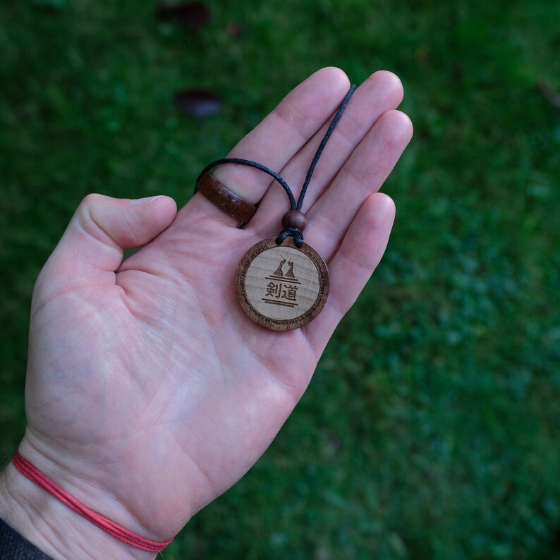 Puede incluir: Un collar con un colgante de madera y un cord&oacute;n negro. El colgante es redondo y presenta un dise&ntilde;o de dos figuras con la palabra "剣道" en caracteres japoneses.