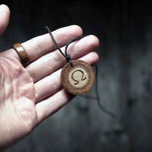 May include: A wooden pendant necklace with a black cord. The pendant is round and features a carved omega symbol surrounded by a decorative border.
