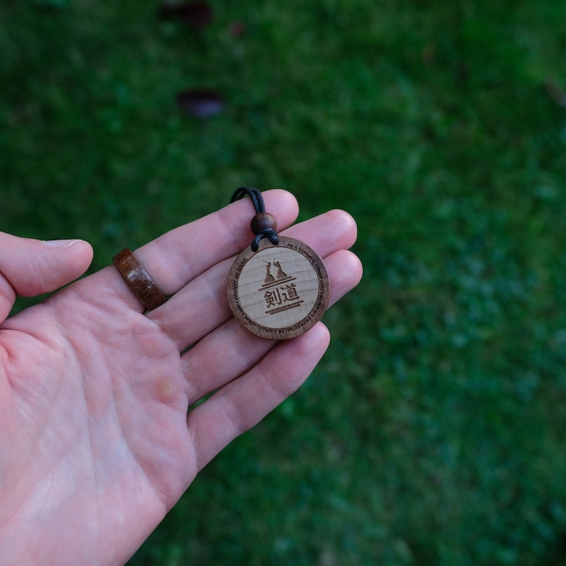 Puede incluir: Un colgante de madera con un cord&oacute;n negro, que presenta una imagen estilizada de dos figuras y el texto "剣道" en caracteres japoneses.